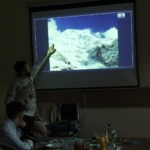 Na pierwszym planie widok ośnieżonych szczytów górskich w Himalajach. Mężczyzna na ekranie projekcyjnym pokazuje widok szczytów górskich w Himalajach na zdjęciu z 1979 roku. 